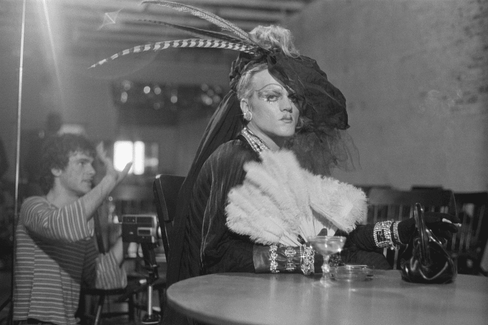 A black-and-white portrait of a person wearing a dramatic feathered headpiece while seated indoors