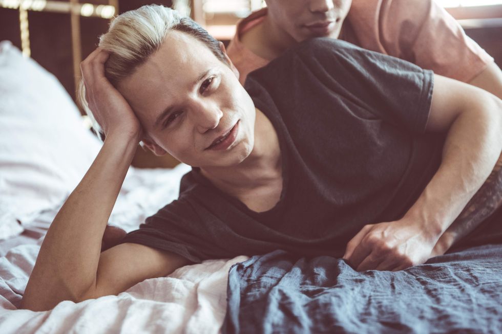 A blond man being spooned in bed by another man
