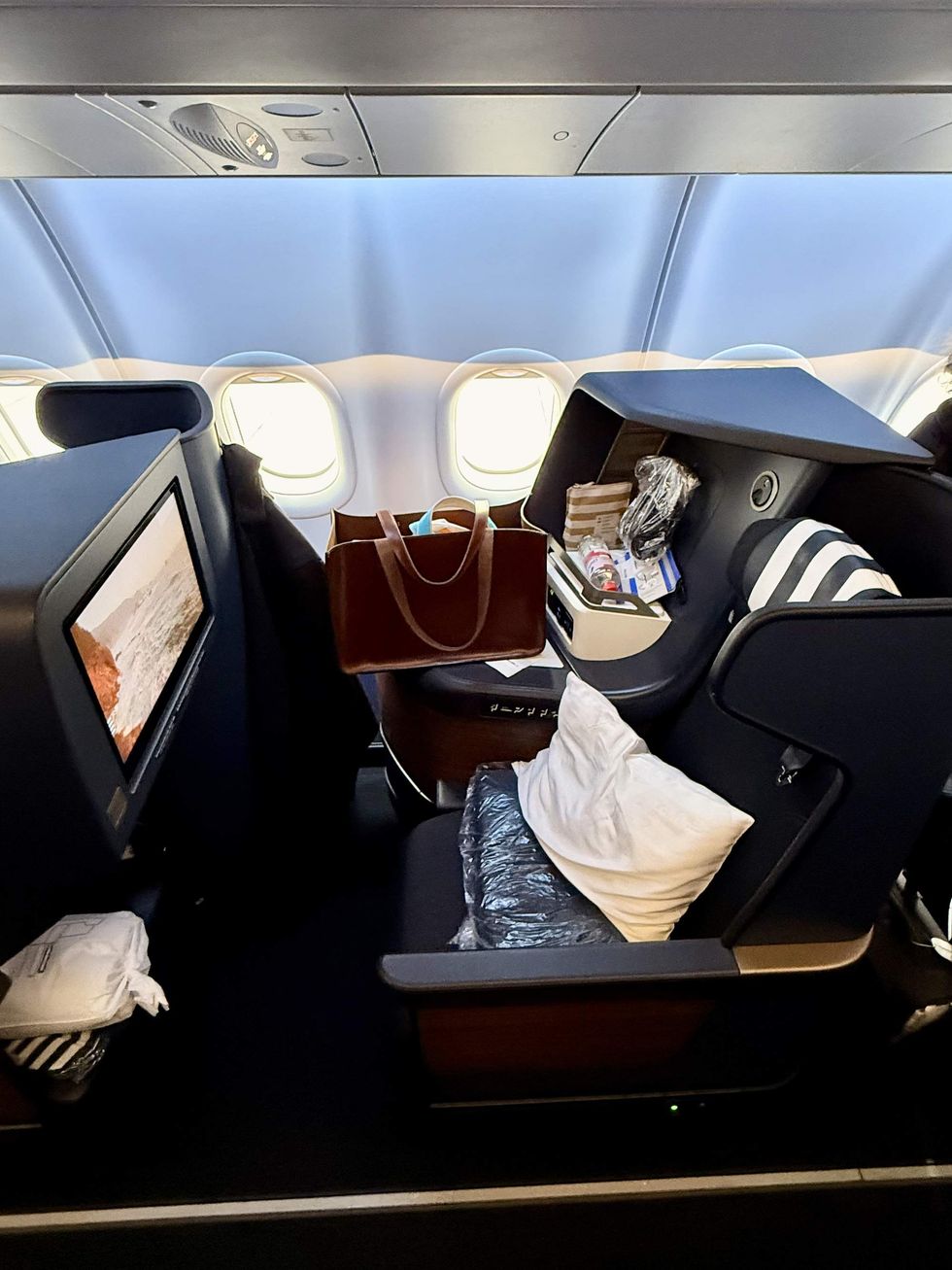 A business class airplane seat with a brown leather bag and coat