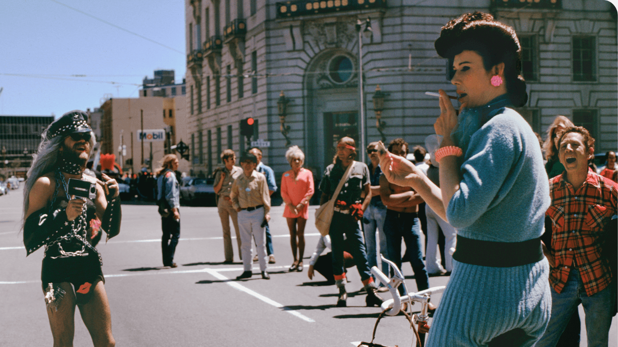 A colorful crowd gathers on a San Francisco street with two people in the foreground facing each other. A person on a bike is smoking a cigarette, the other is filming in a leather outfit