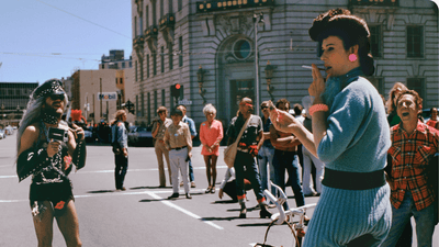A colorful crowd gathers on a San Francisco street with two people in the foreground facing each other. A person on a bike is smoking a cigarette, the other is filming in a leather outfit