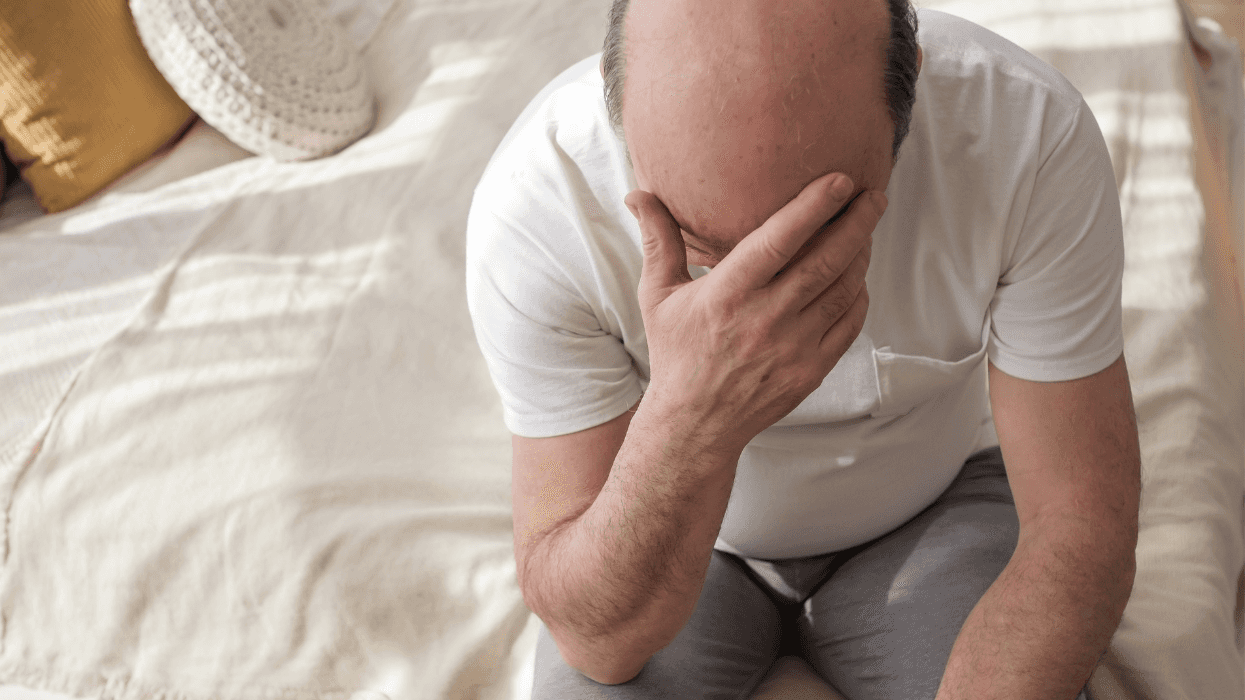 A distressed older man sits on the edge of a bed with his face buried in one hand, appearing overwhelmed and deep in thought