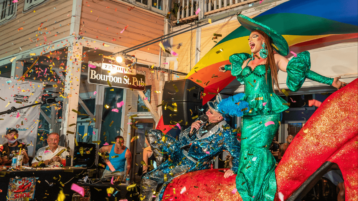 A drag performer in a sparkling green outfit and matching hat celebrates atop a giant red high heel as colorful confetti fills the stage.