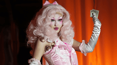 A drag performer with voluminous pastel pink hair and dramatic pink makeup poses on stage in a pink corset dress and long white gloves, holding cash in one hand against a warm orange curtain backdrop.