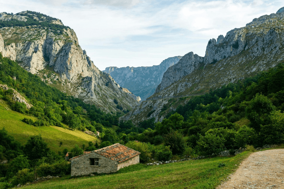 A lush green valley surrounded by dramatic rocky mountains, with a small stone building in the foreground.