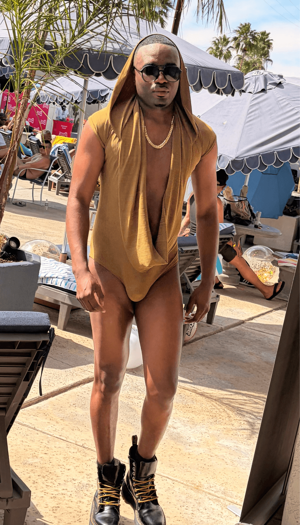 A model wearing a gold hooded bodysuit, sunglasses, and black boots poses poolside during NoRal Apparel\u2019s fashion showcase at SunLit Men of Color Retreat in Palm Springs