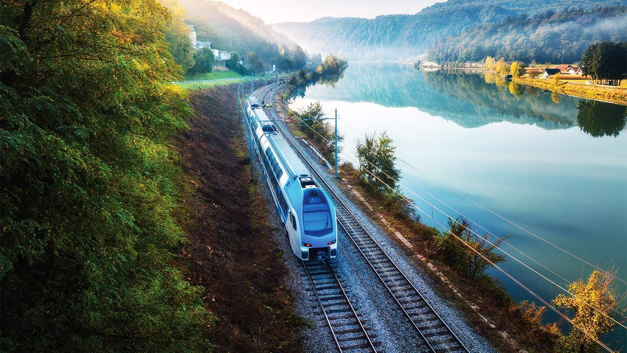 a modern train traveling alongside a river through a green hilly landscape