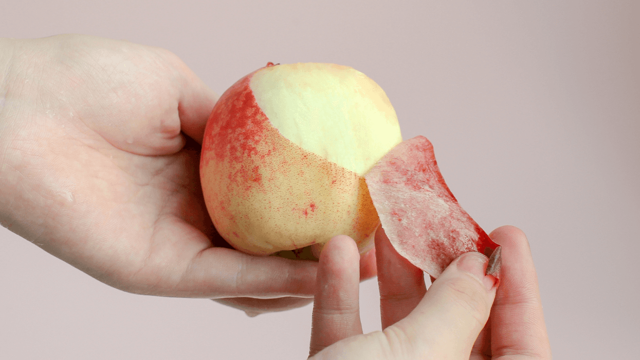 A person peeling the skin off a peach, exposing the smooth fruit underneath while holding it in their hands