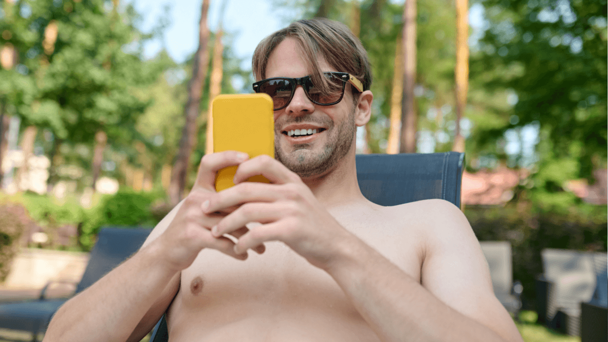 A shirtless man wearing sunglasses smiles while looking at his phone outdoors.