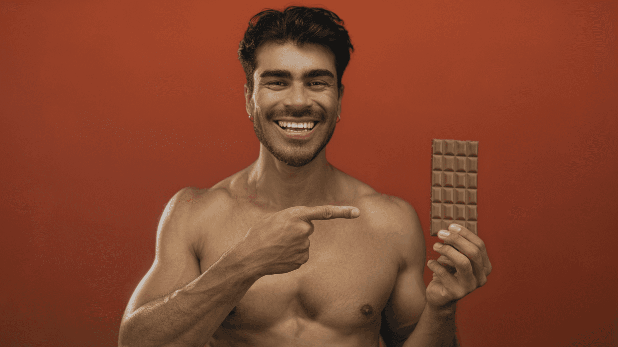 A smiling shirtless man points to a chocolate bar while posing against a burnt orange background