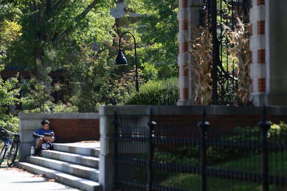 A student sits on the stairs of a red brick building and reads a book. A bike is parked beside them, resting on a wall.