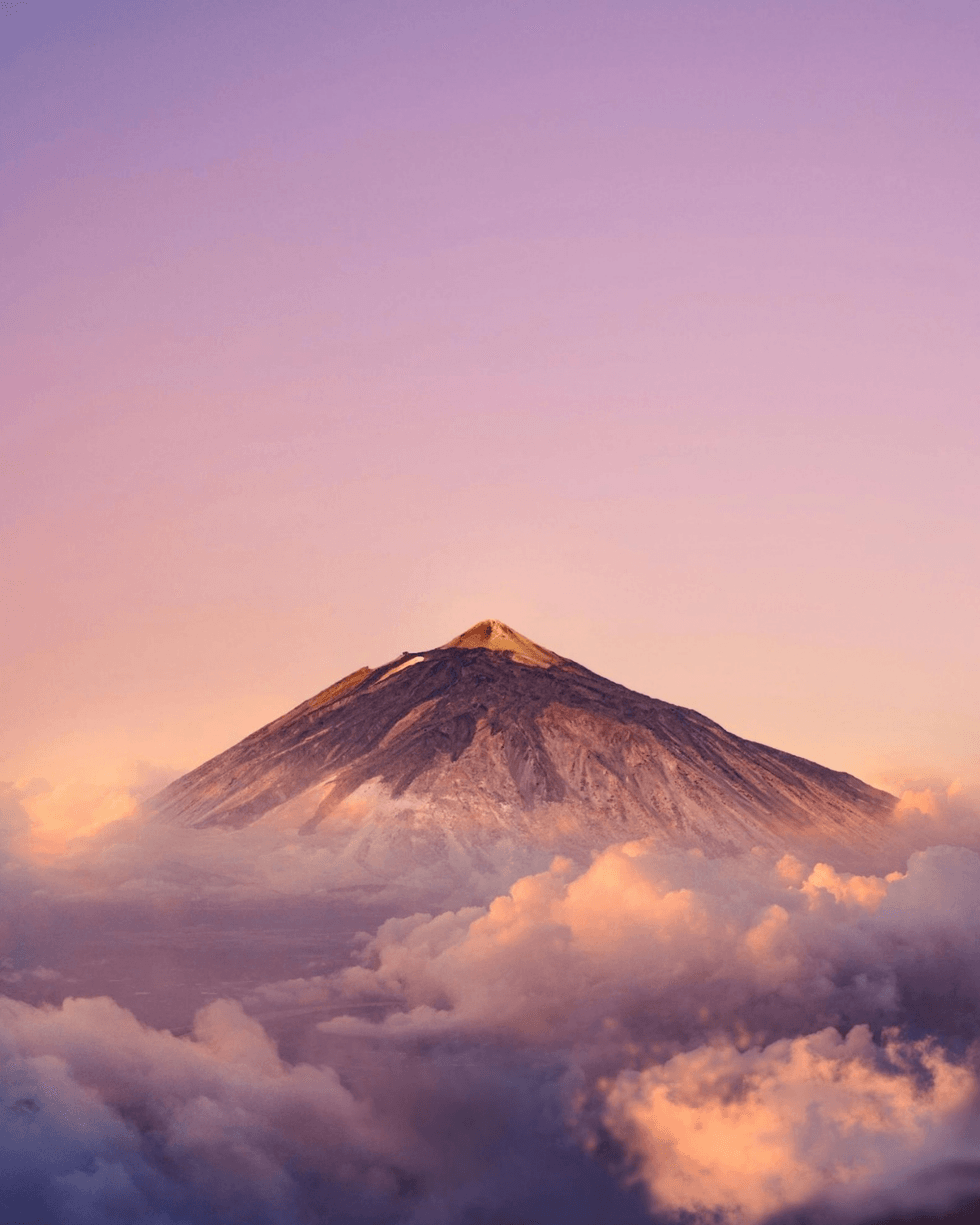 A volcanic peak rising above a layer of clouds beneath a pink and purple sky.