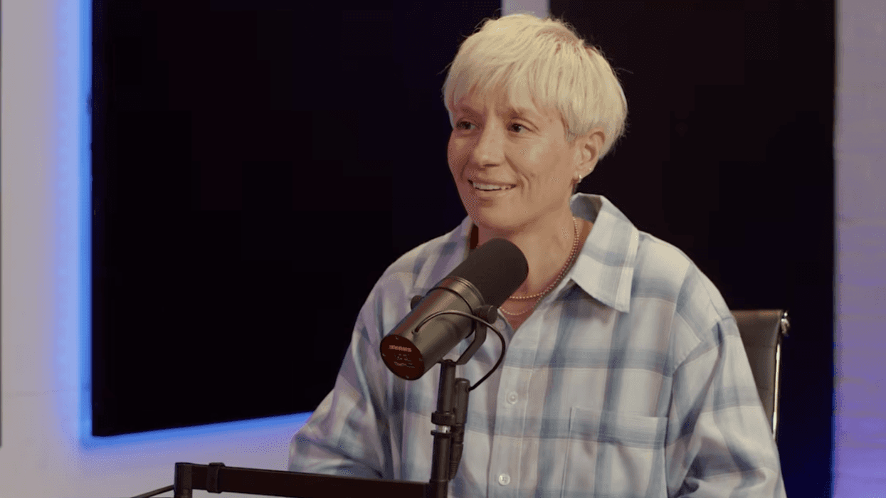 A woman with blonde hair and a blue and white checkered shirt sits in front of a black microphone during an interview for a podcast.