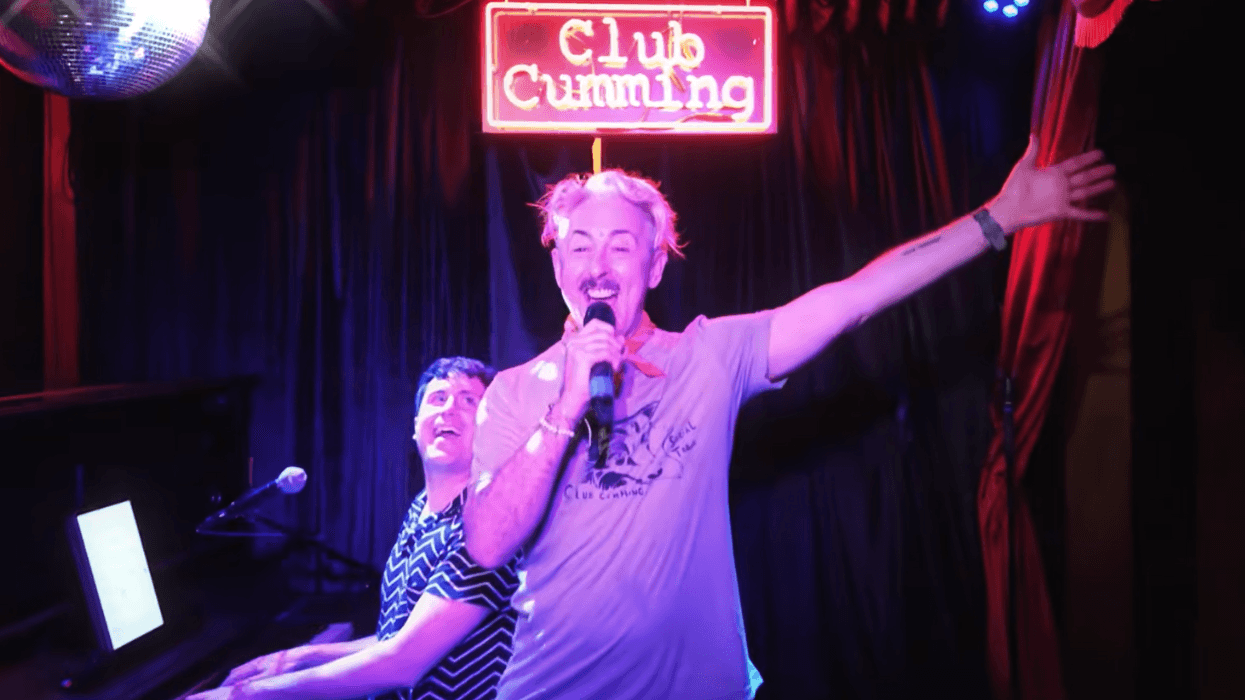 Alan Cumming speaks onstage at Club Cumming under a neon sign while a pianist sits beside him