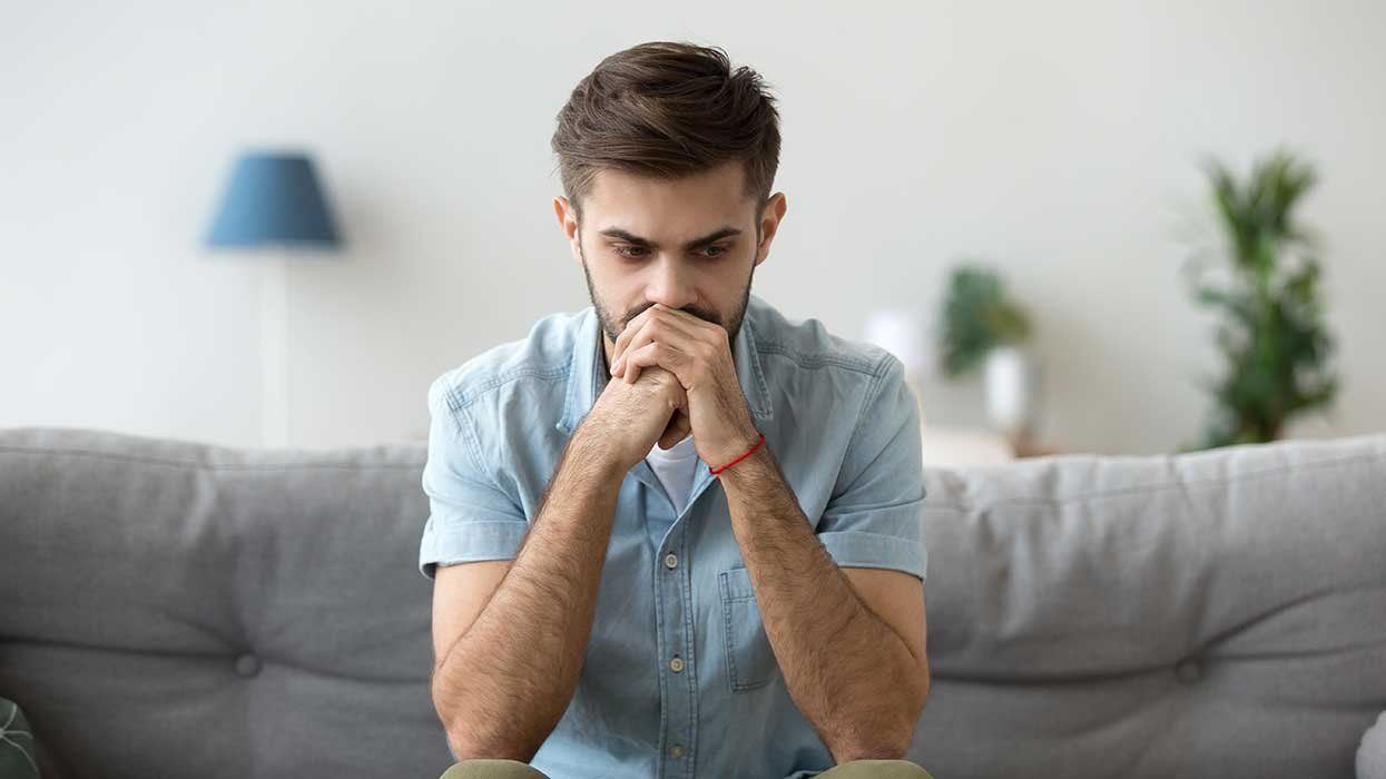 Contemplating young millennial serious man sitting on couch alone home