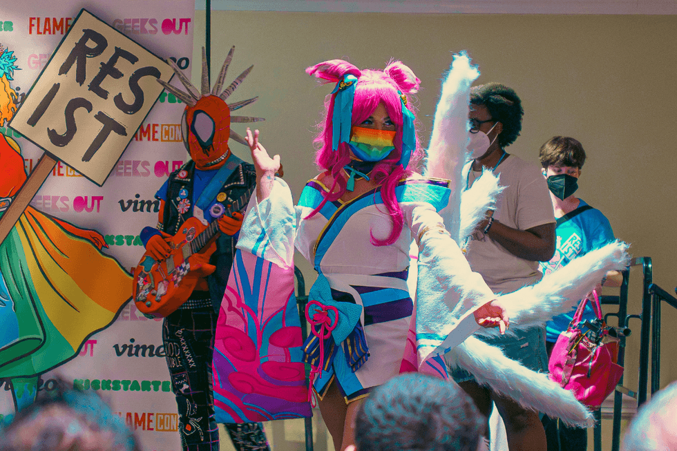 Cosplayer in a colorful anime-inspired costume and rainbow mask poses onstage during a Flame Con event