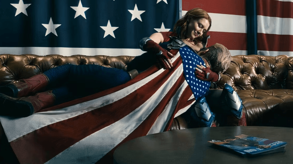 Firecracker embraces Homelander on a couch beneath a large American flag in a scene from The Boys.
