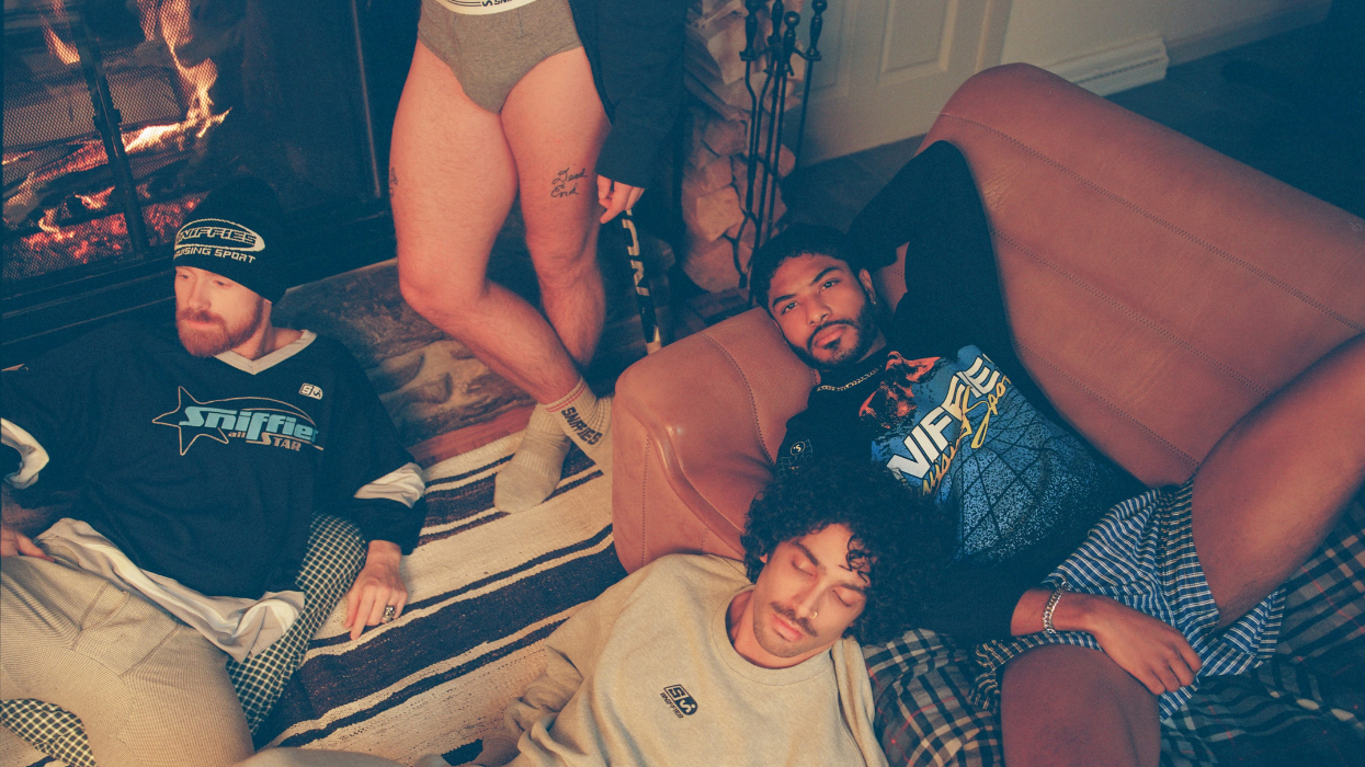 Four men in a living room, two are on the floor and one is on the couch, another is standing by the fireplace