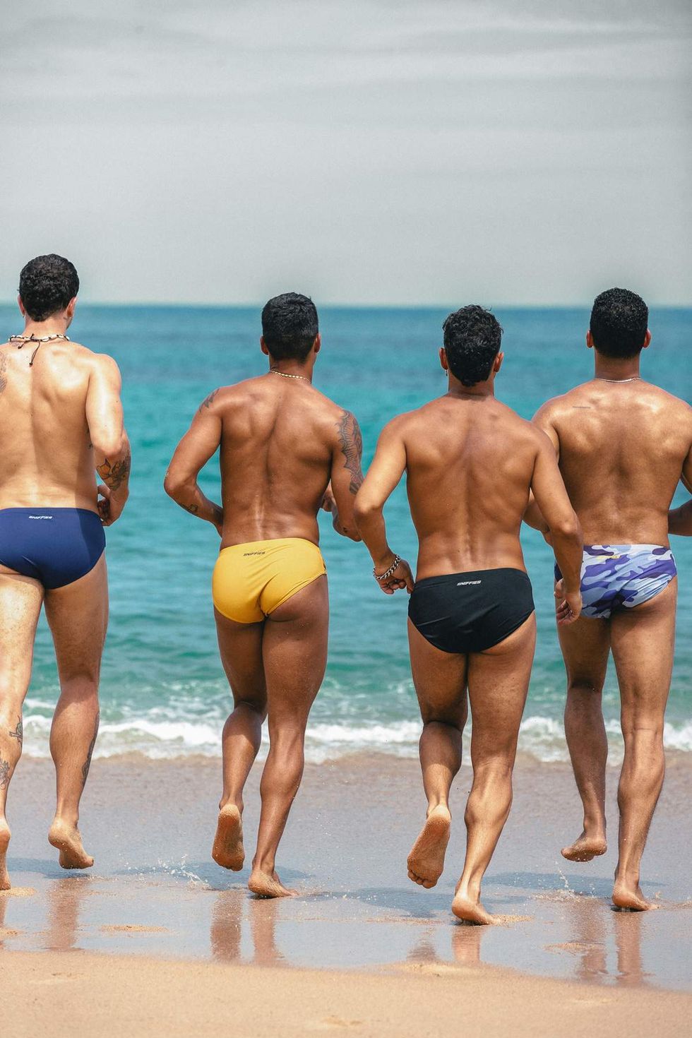 Four models wearing Sniffies swim briefs run toward the ocean during the Sniffies Summer Swim Collection campaign in Puerto Rico