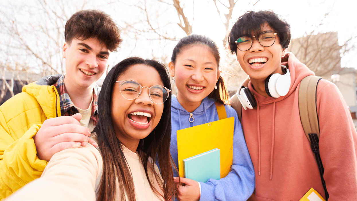 Four smiling students with backpacks and school supplies pose together outdoors.