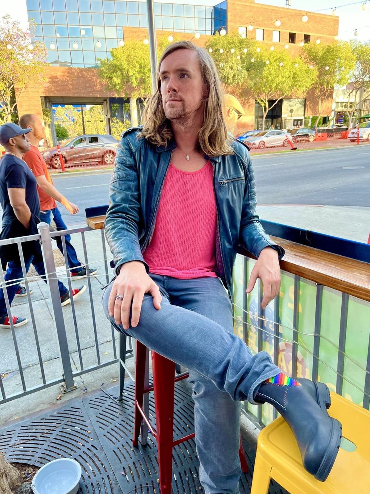 Genderfluid individual sitting outside a bar in Los Angles, their legs are crossed and they are wearing rainbow boots
