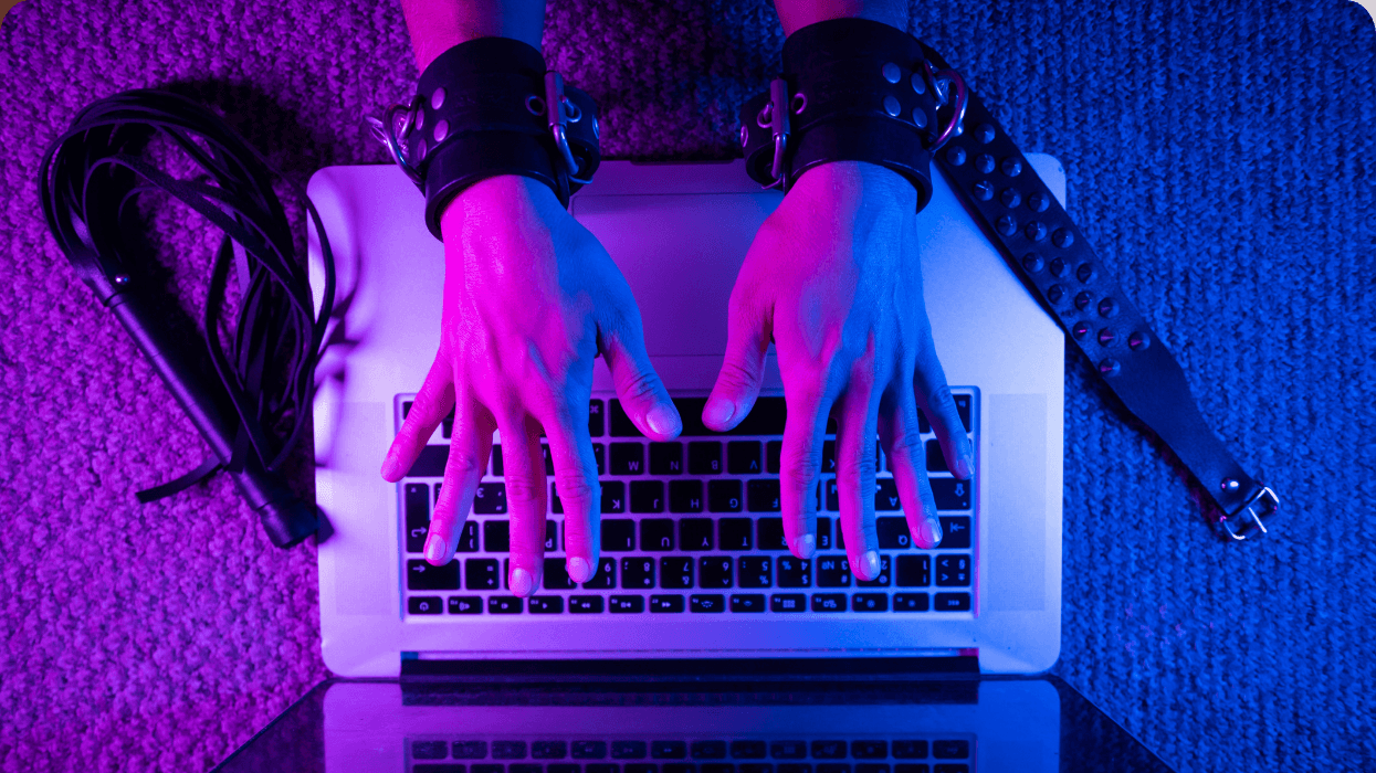 Hands wearing leather wrist restraints type on a laptop beside BDSM gear under pink and blue lighting.