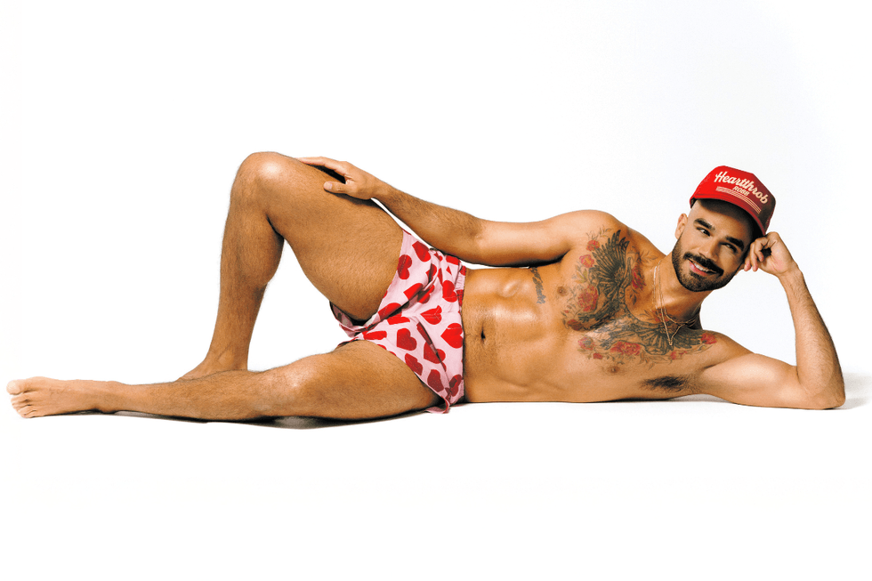 Heartthrob Robb reclining shirtless in a red cap and heart-print shorts, showing chest tattoos.