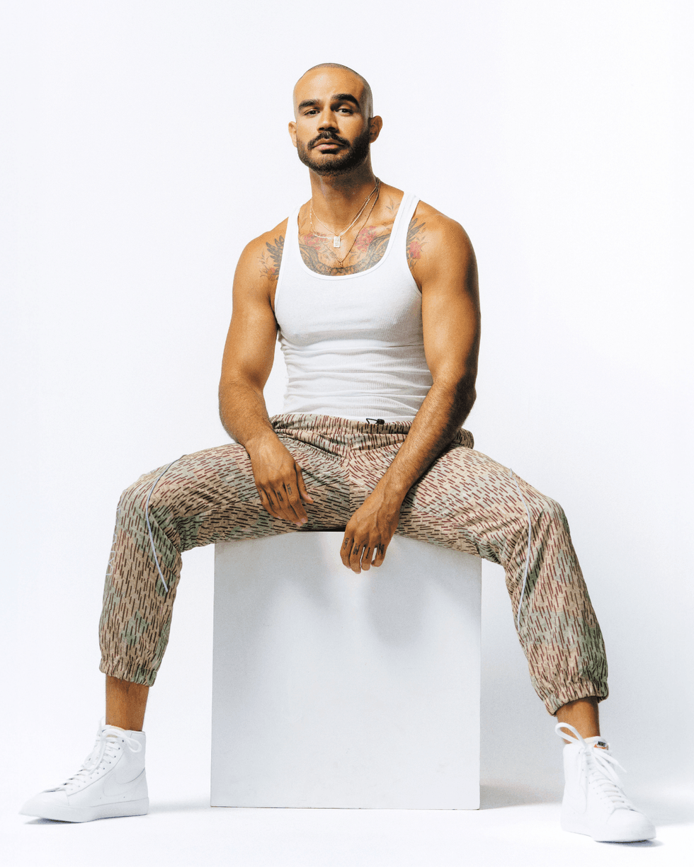 Heartthrob Robb sitting on a white cube wearing a white tank top, patterned pants, and white sneakers, showing chest tattoos.