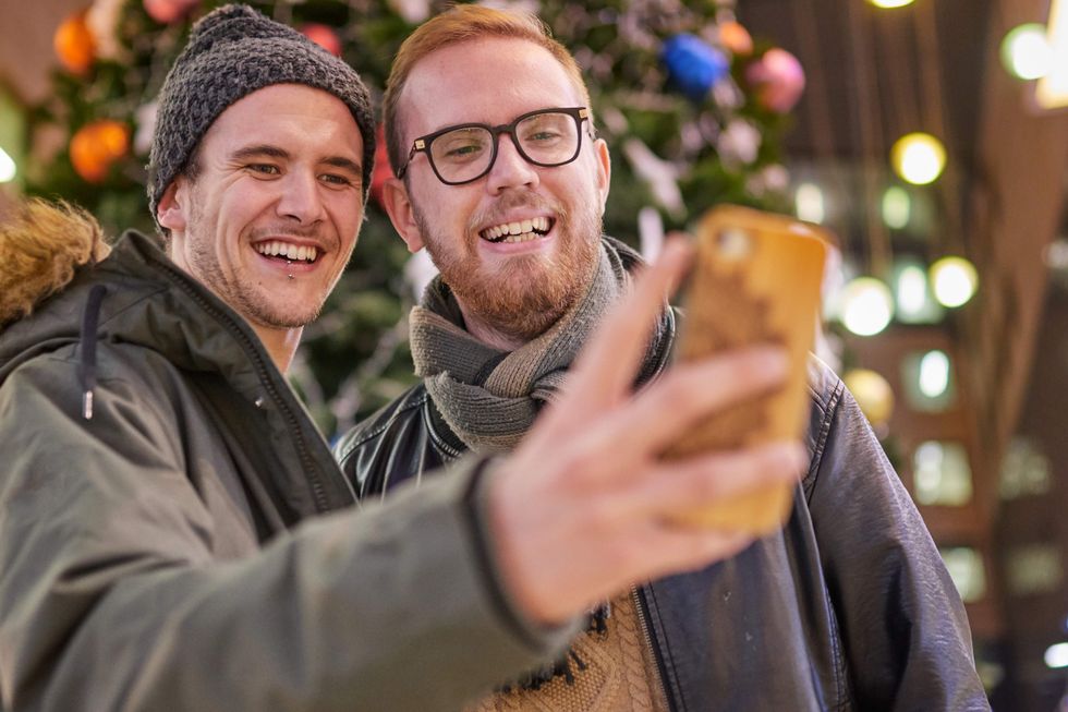 homosexual couple of boys, taking a selfie, are smiling, in front of a Christmas tree, inside a shopping mall