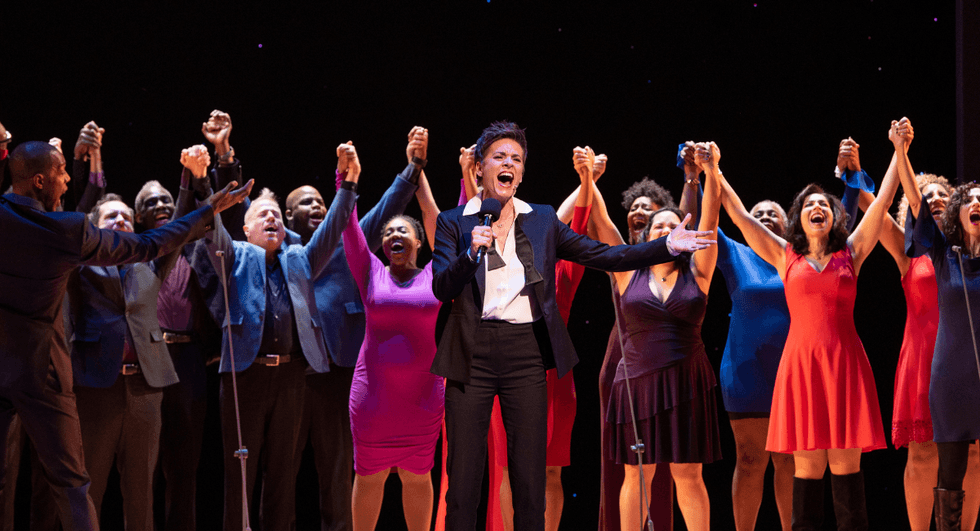 Jenn Colella holds a microphone while the Broadway Backwards cast stands behind her onstage with raised hands.