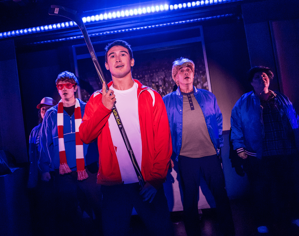 Jimin Moon stands center stage holding a hockey stick while performers surround them in Heated Rivalry: The Unauthorized Musical Parody.