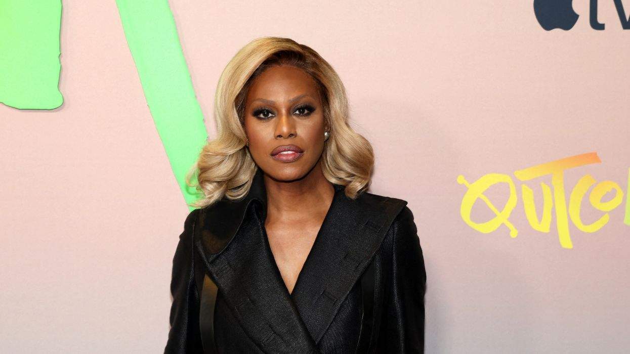 Laverne Cox with styled blonde hair in a black coat at an event with colorful graphics behind.