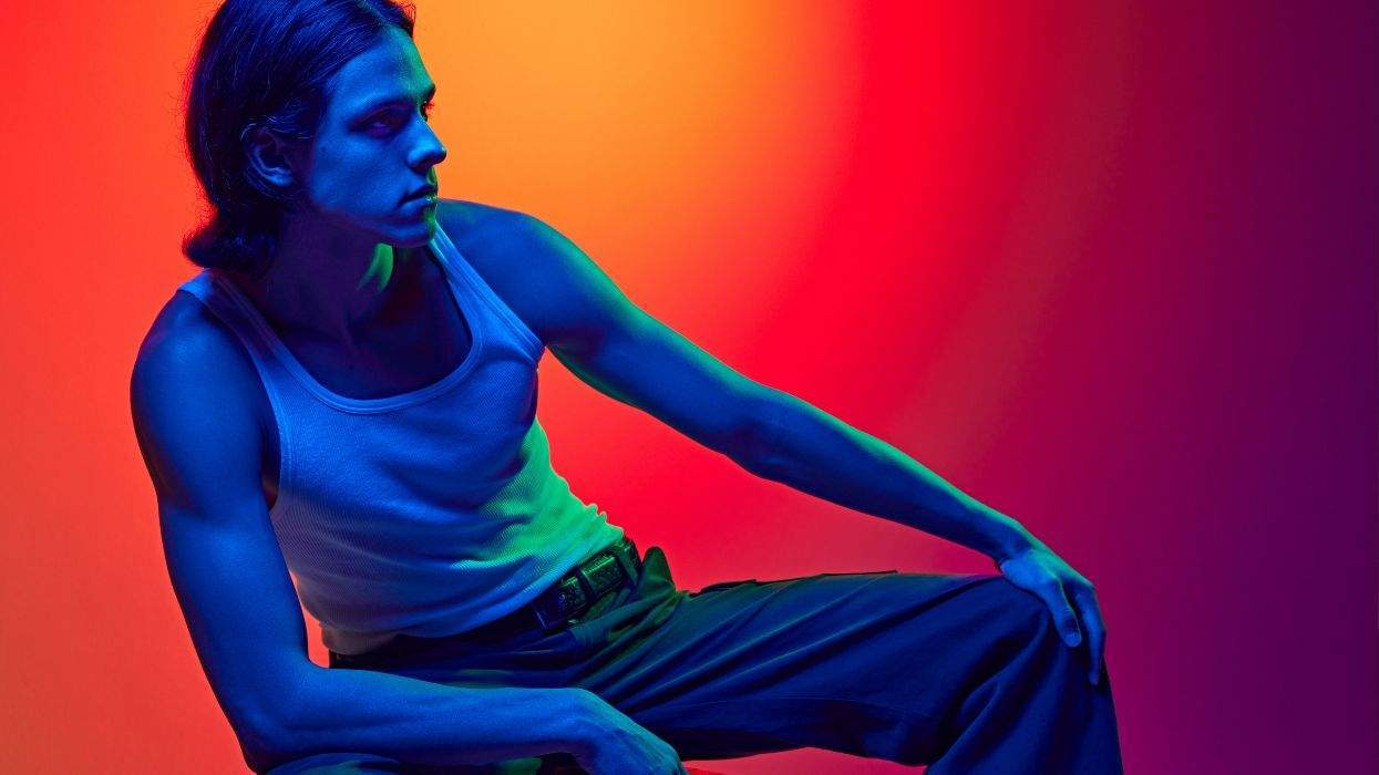 Man in a blue light, wearing a tank top, poses against a vibrant orange-red background.