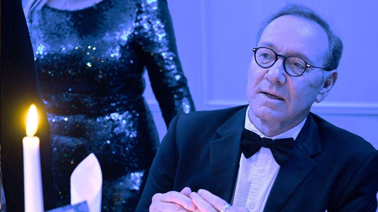 Man in tuxedo and glasses sits at a candle-lit table, with a woman in sequined dress nearby.