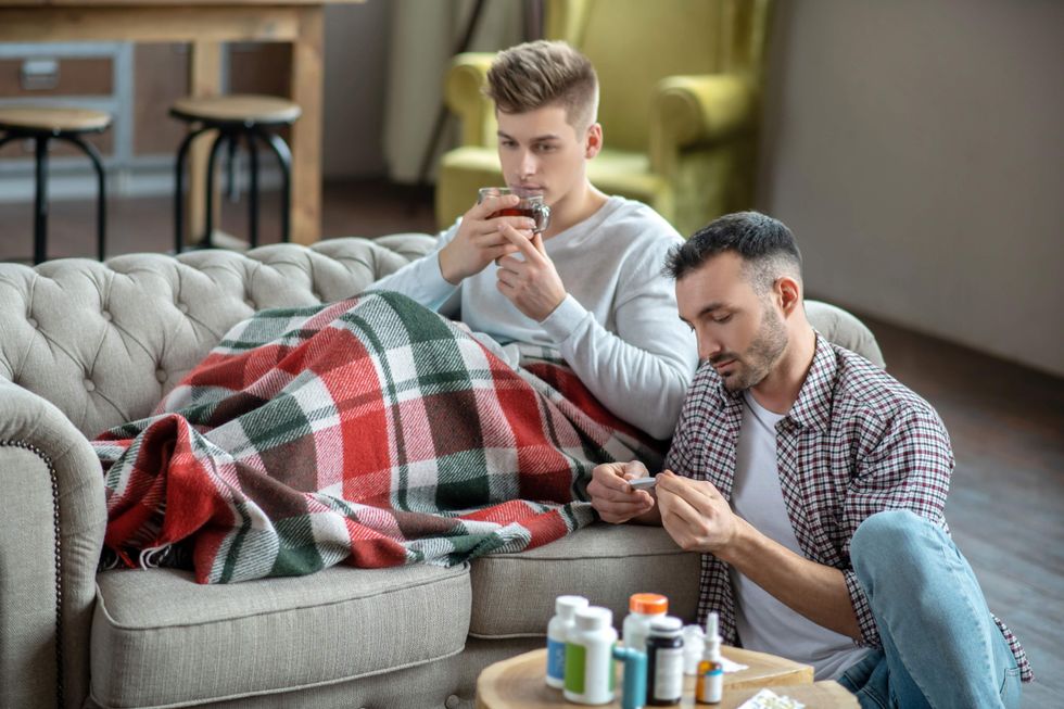 Man on a couch drinking tea and a second man sitting on the floor holding a thermometer