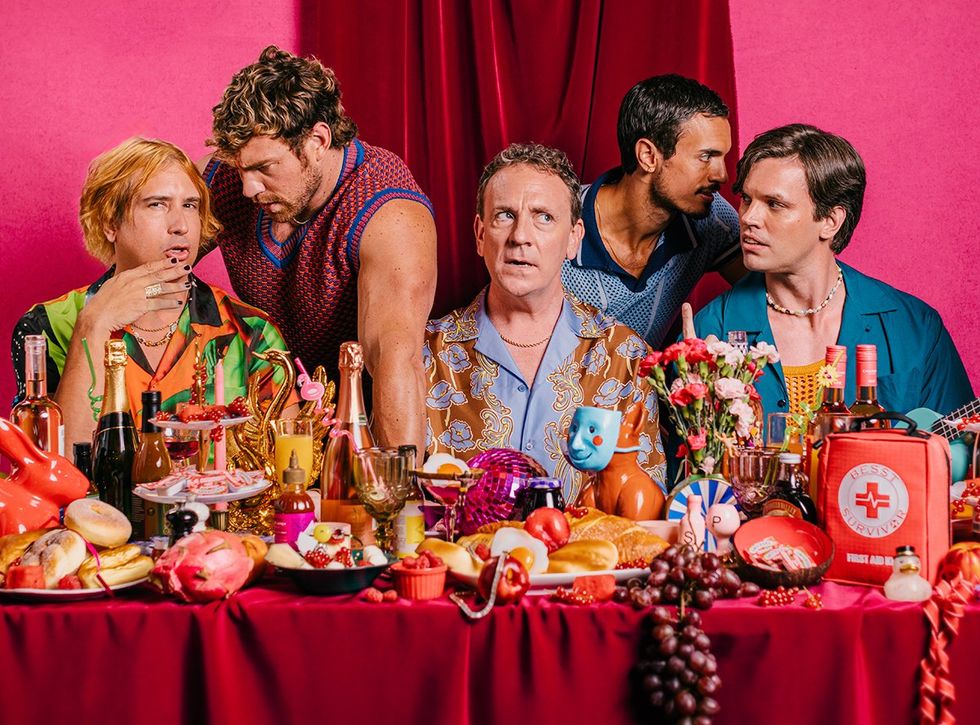 MESSY WHITE GAYS show Drew Droege at the center surrounded by cast whispering to each other all at a long feast table full of lavish colorful stage props