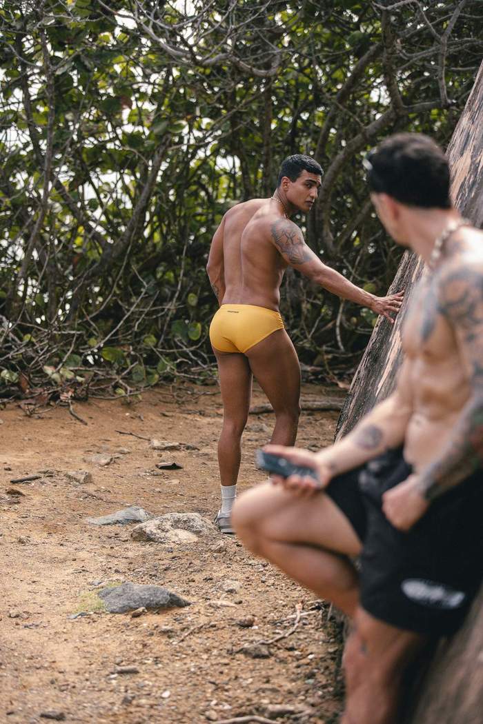 Models wearing Sniffies swimwear share a flirtatious moment along a coastal path during the Sniffies Summer Swim Collection campaign in Puerto Rico.