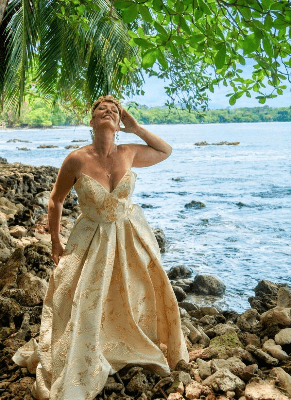 Nina Rose Fischer poses on a rocky beach in Costa Rica wearing a gold strapless wedding gown