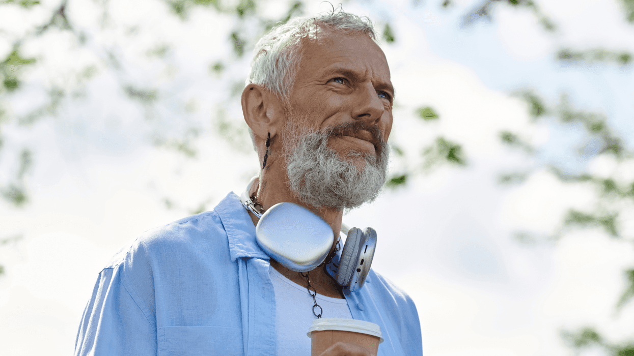 Older gray-haired man wearing headphones and holding a coffee outdoors