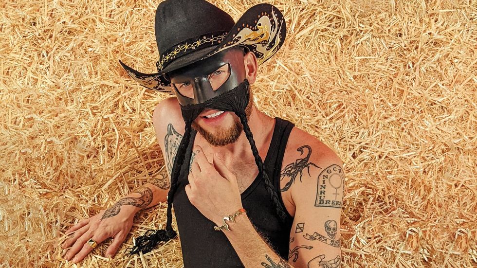 ​Orville Peck laying in a haystack smiling at the camera