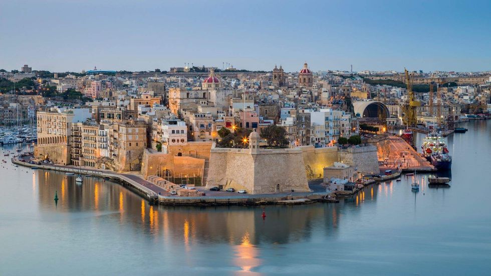 Panoramic view of Valletta, Malta, at dusk with historic buildings, fortified walls, and reflections across the harbor water