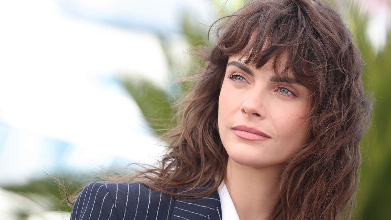 Person with long hair in a pinstripe suit looks thoughtfully into the distance.