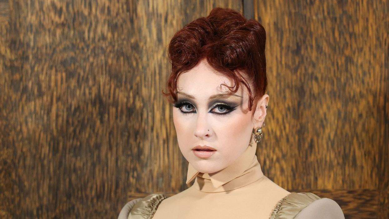 Person with styled red hair, dramatic makeup, in front of a wooden background.