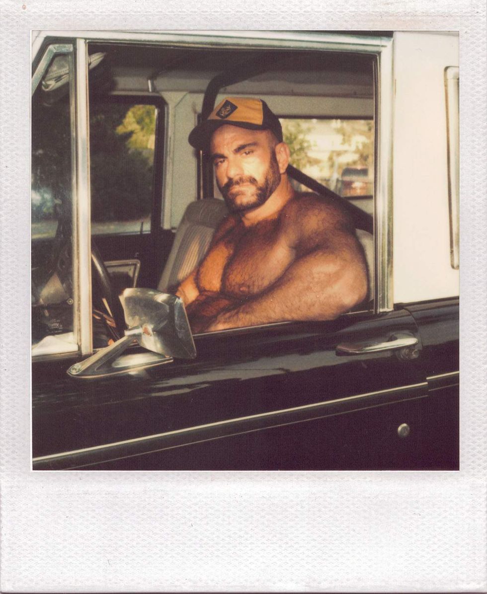 Polaroid of a bearded, shirtless man wearing a baseball cap while sitting in the driver\u2019s seat of a vintage pickup truck, looking toward the camera through the open window. Image from Michael Alago\u2019s book Polaroid Encounters