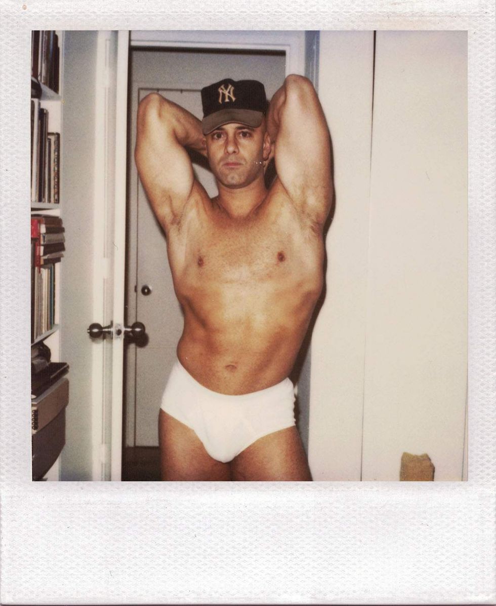 Polaroid of a man wearing white briefs and a New York Yankees baseball cap, posing indoors with both hands behind his head while standing in a doorway beside a bookshelf. Image from Michael Alago\u2019s book Polaroid Encounters