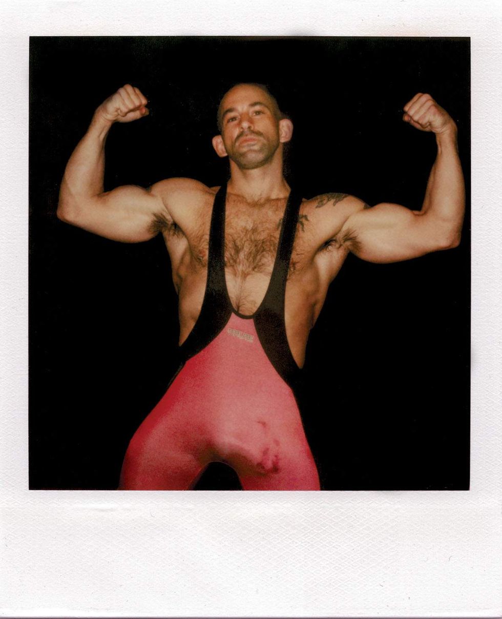 Polaroid of a muscular man in a red wrestling singlet flexing both arms against a black background, looking directly at the camera