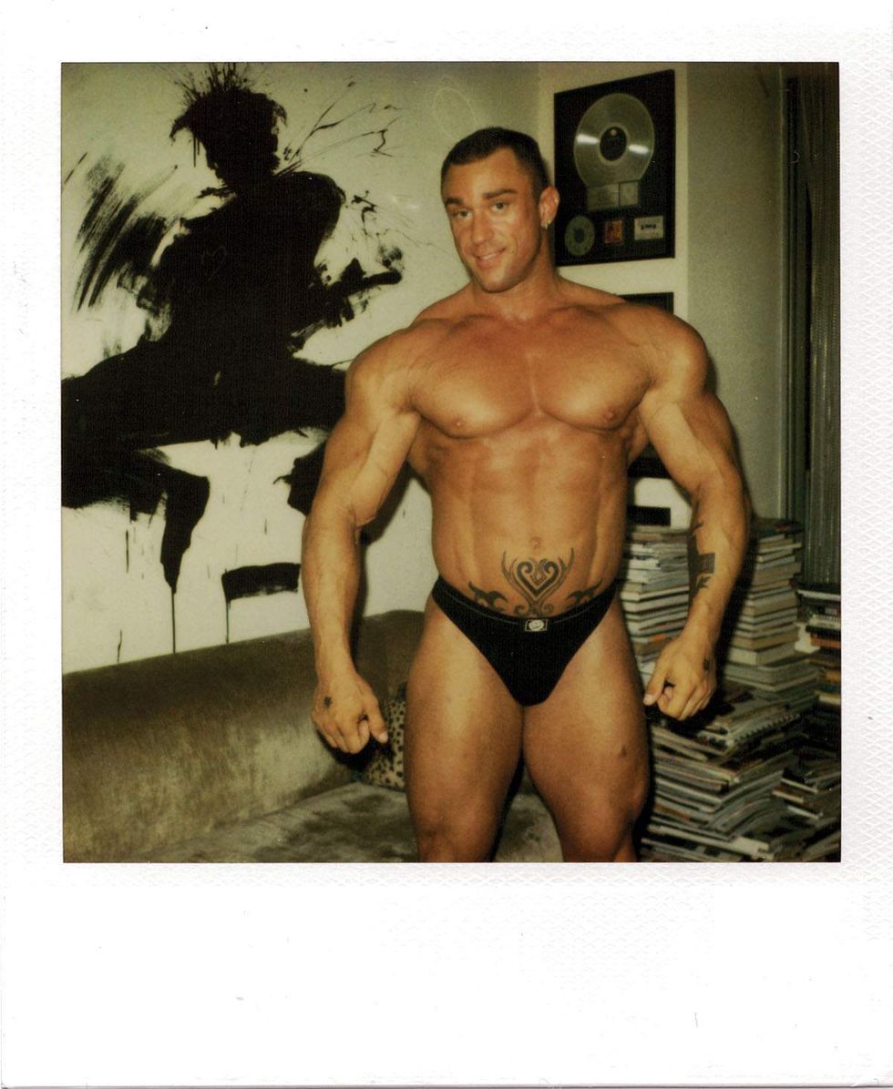 Polaroid of a muscular man wearing black briefs and posing indoors beside a couch and stacks of magazines, with framed artwork and a silver record plaque hanging on the wall behind him. Image from Michael Alago\u2019s book Polaroid Encounters