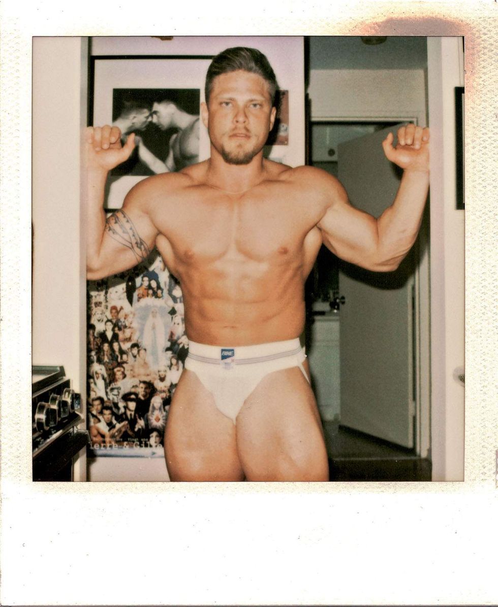 Polaroid of a muscular man wearing white briefs and flexing both arms while standing in a doorway inside an apartment. Posters and framed artwork are visible behind him. Image from Michael Alago\u2019s book Polaroid Encounters