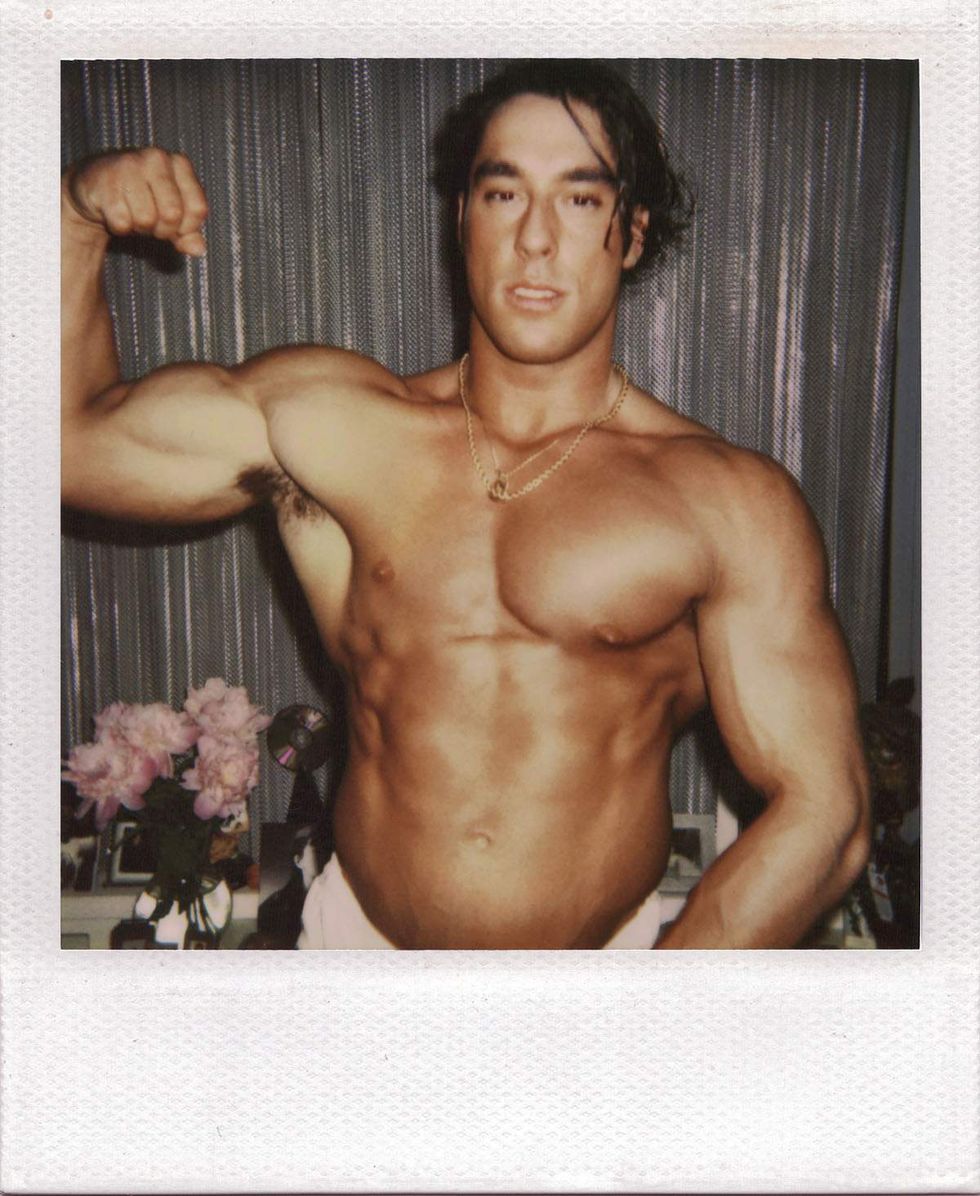 Polaroid of a muscular man with dark hair flexing one arm while posing shirtless indoors, wearing a gold chain necklace in front of metallic curtains and a table with pink flowers. Image from Michael Alago\u2019s book Polaroid Encounters
