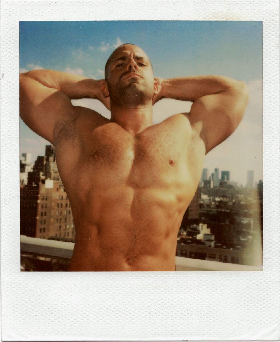 Polaroid of a shirtless muscular man posing on a rooftop with his hands clasped behind his head, standing against a sunlit city skyline and blue sky. Image from Michael Alago\u2019s book Polaroid Encounters